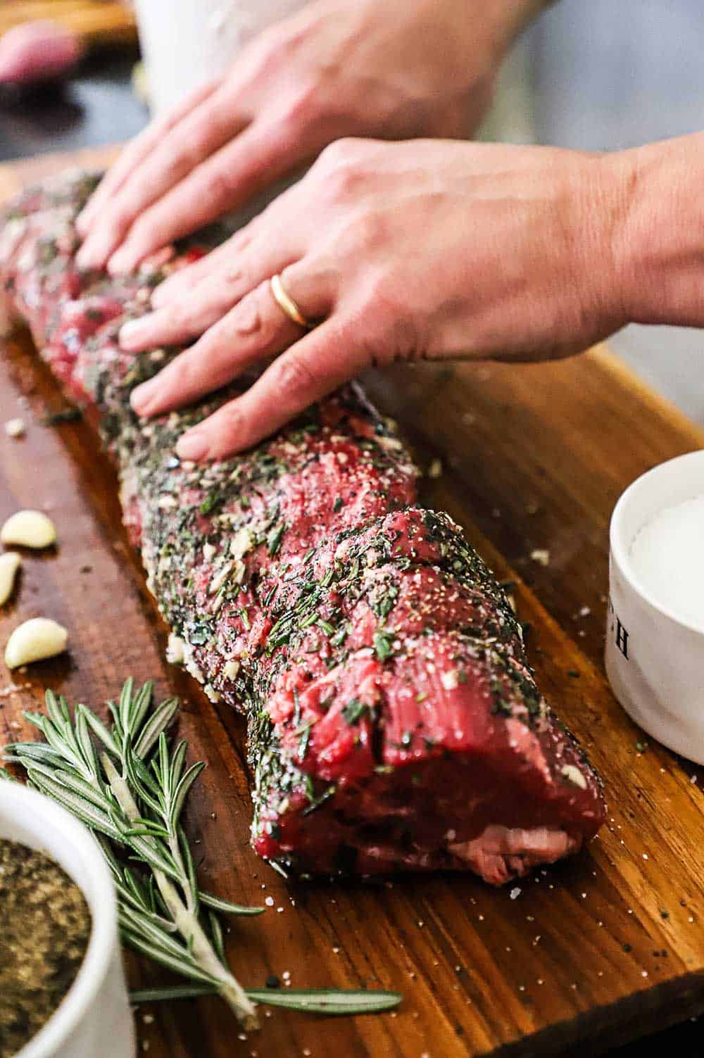 A person rubbing chopped herbs, garlic, salt, and pepper into a large uncooked beef tenderloin. 