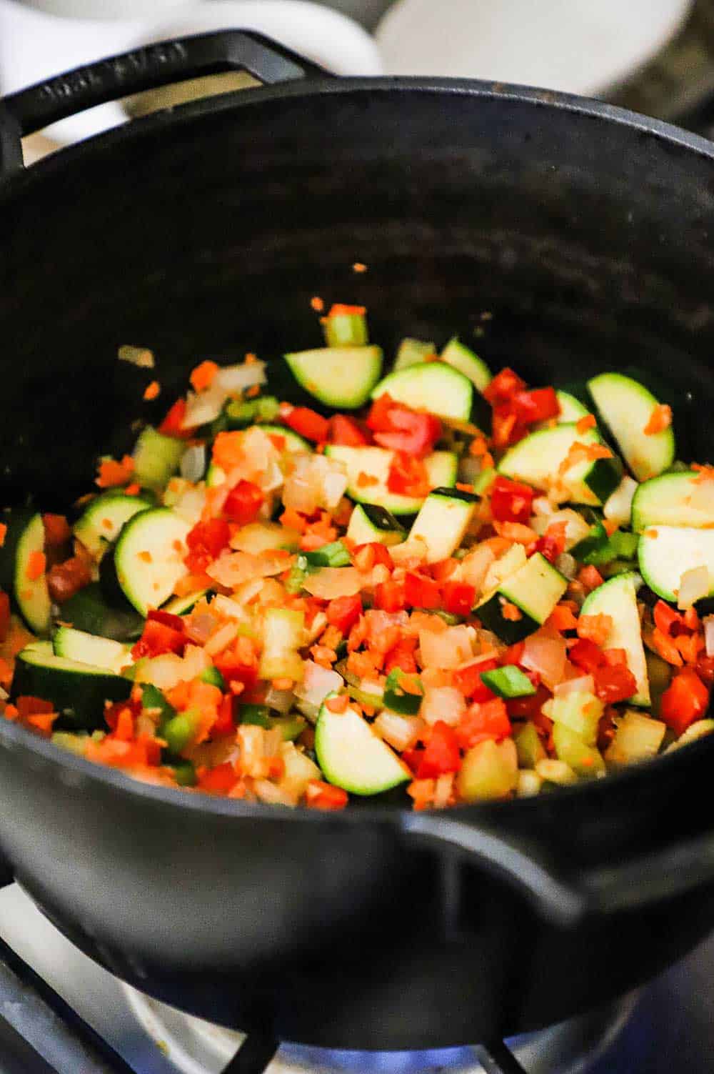 A mixture of chopped zucchini, carrots, onion, and jalapeño being sautéed in a large cast-iron pot.