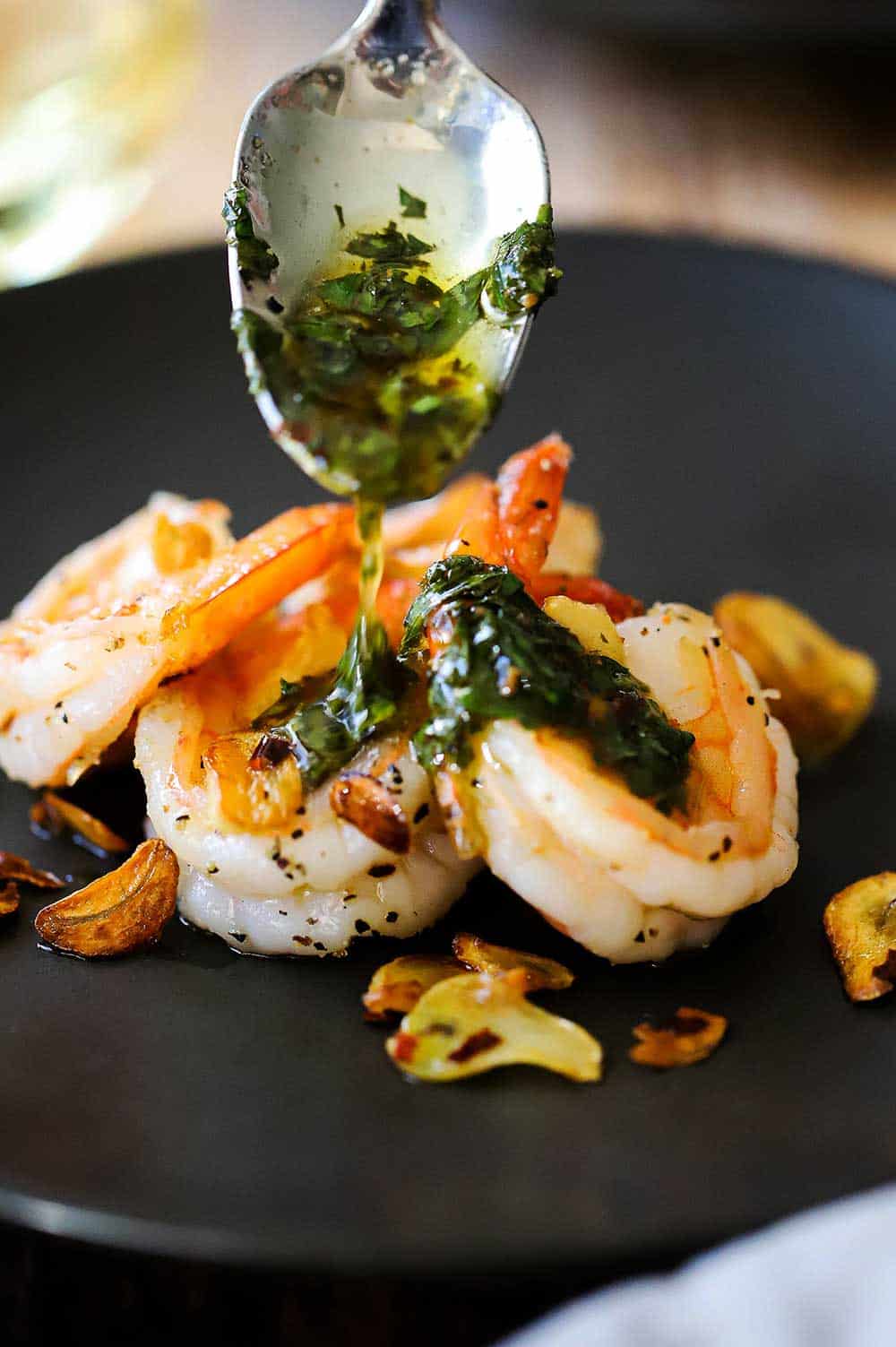 A white wine and parsley sauce being poured over garlic shrimp on a small dark grey appetizer plate. 