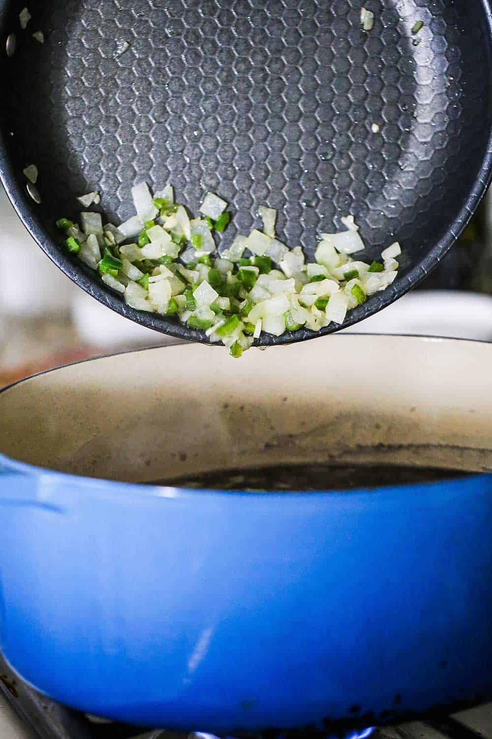 Sautéed chopped onions and jalapeños being transferred from a non-stick skillet into a large blue Dutch oven filled with simmering black beans.