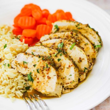 A white dinner plate filled with 5 slices of cut baked honey mustard chicken sitting next to sliced carrots and rice pilaf also on the plate.
