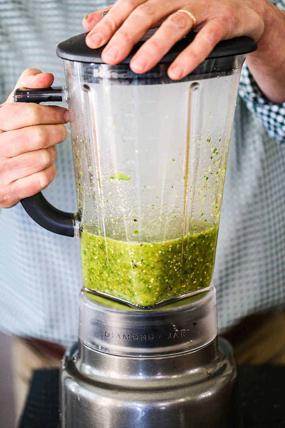 A person holding onto the lid of a blender that is filled with puréed salsa verde.