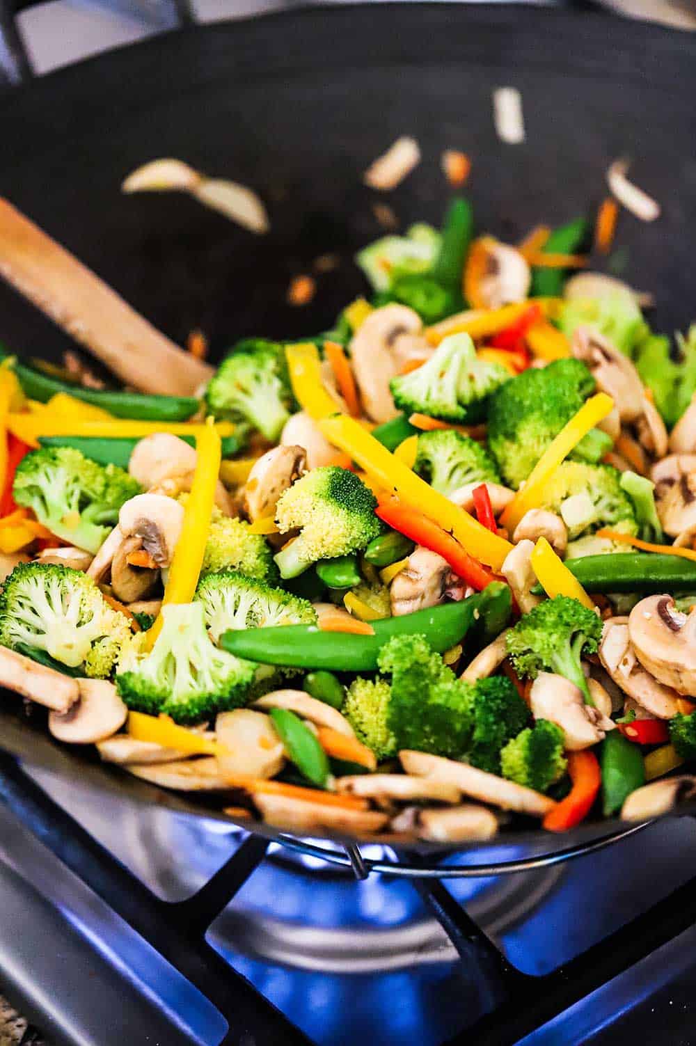 A large black wok over a gas flame on a stove with vegetable stir-fry cooking inside it. 