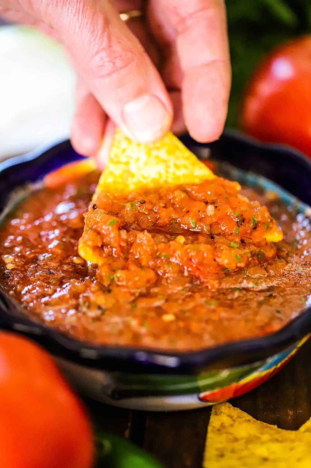 A person's hand dipping a corn tortilla chip into a small festive bowl filled with roasted tomato salsa.