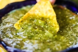 A person dipping a corn tortilla chip into a festive bowl filled with salsa verde.