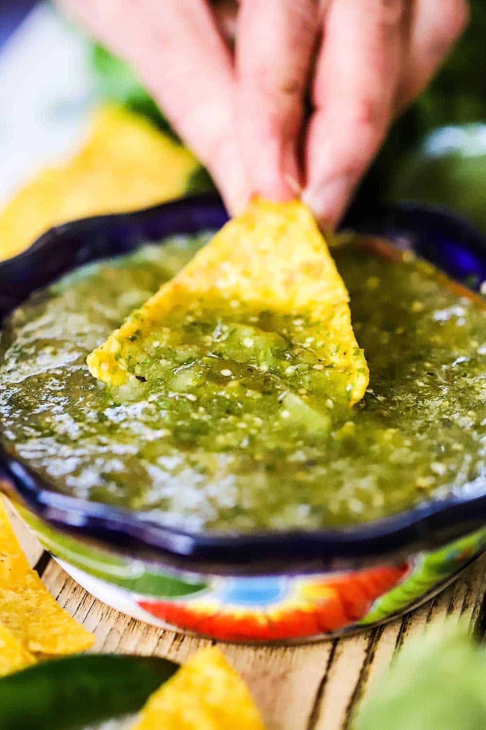 A person dipping a corn tortilla chip into a festive bowl filled with salsa verde.