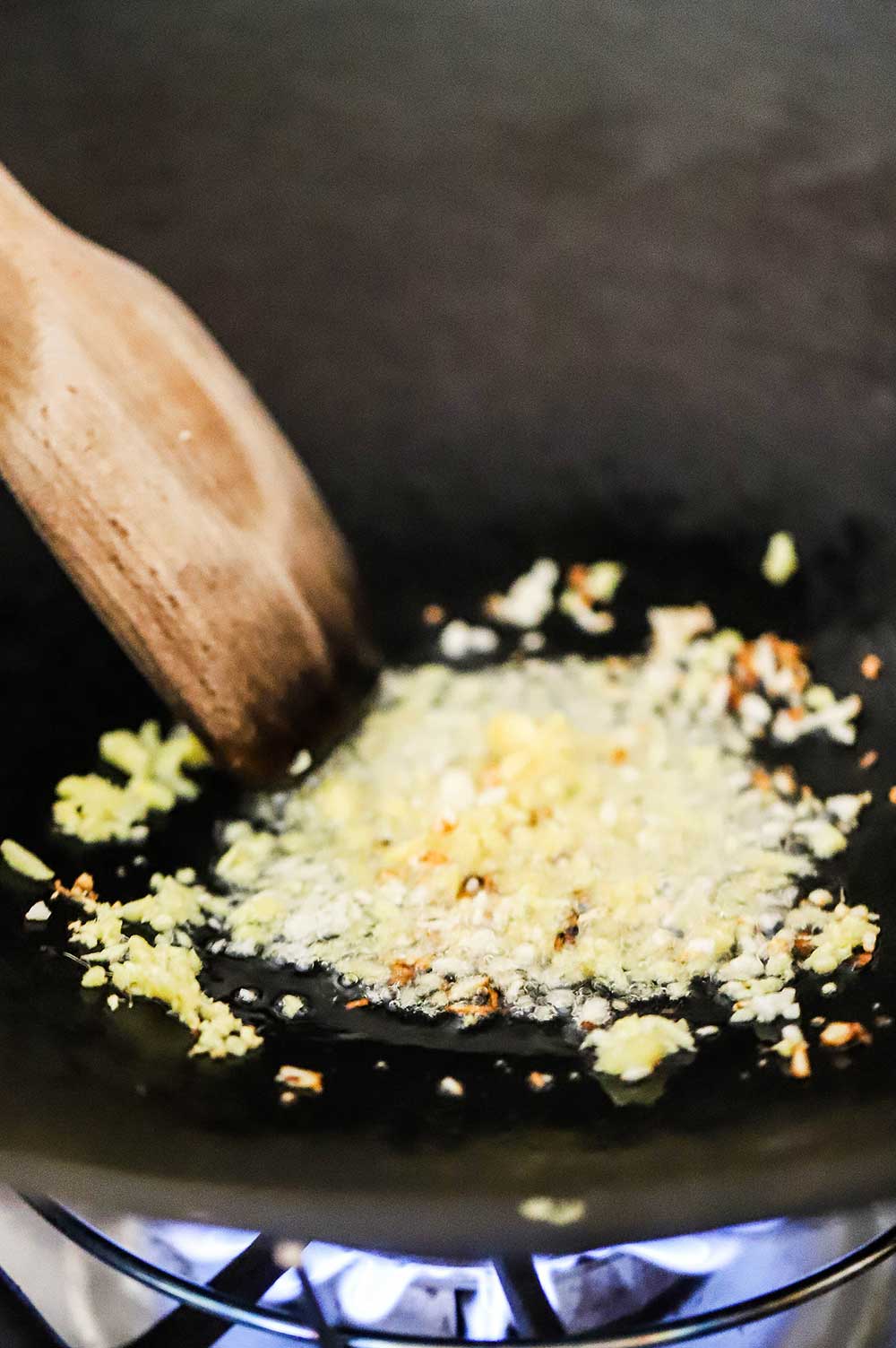 Minced garlic and fresh ginger being stirred fried in a black wok with a wooden spatula. 