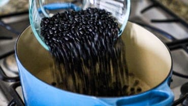 Dried black beans being transferred from a glass bowl into a large oval blue Dutch oven sitting on a stove.