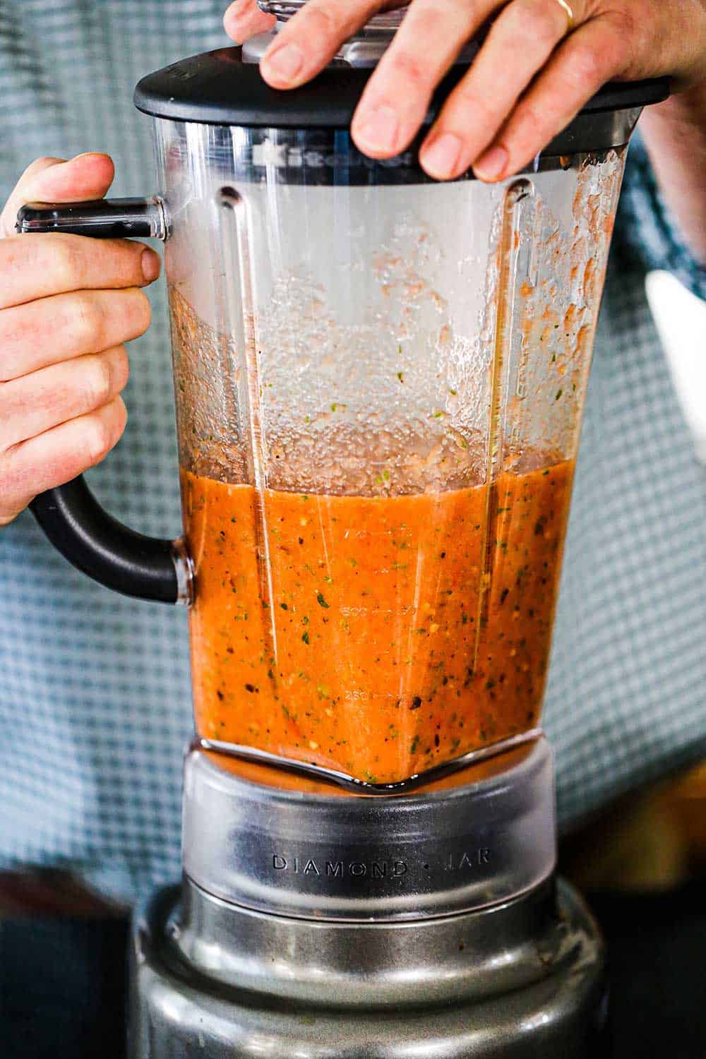 A person holding onto the lid of a blender filled with puréed roasted tomatoes and other vegetables.