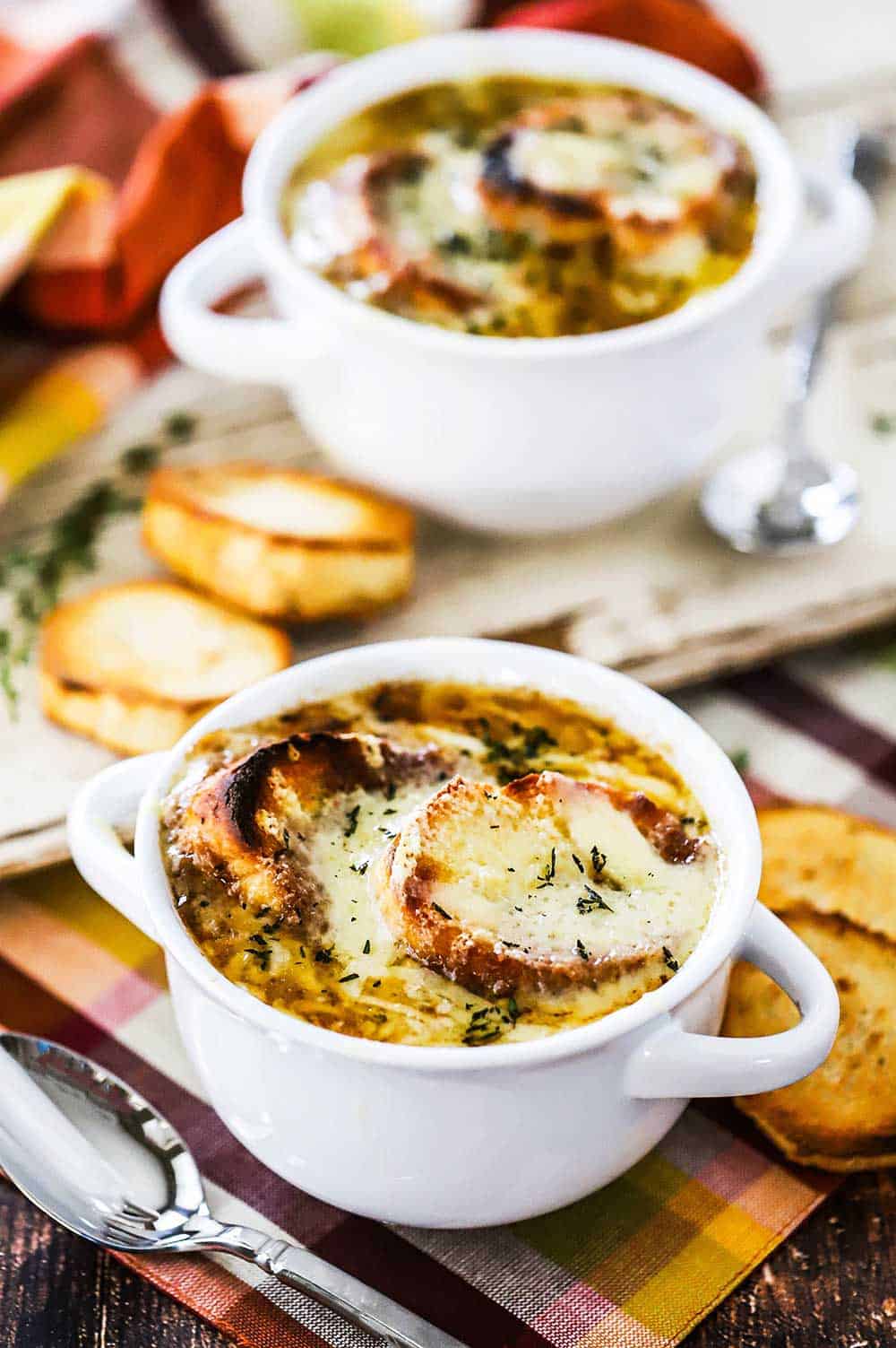 Two white bowls filled with French onion soup with sliced toasted pieces of a baguette and spoons nearby.