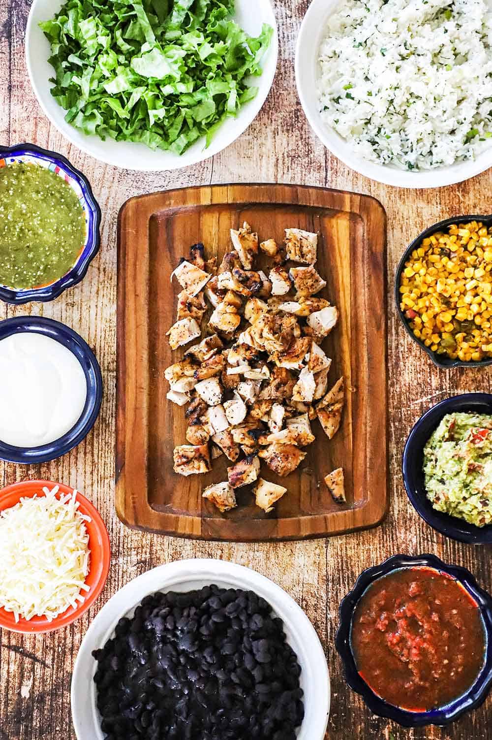 A small wooden cutting board filled with grilled chopped chicken surrounded by bowls of salsa, beans, rice, lettuce, and sour cream.