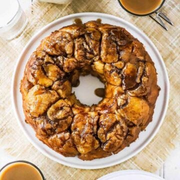 An untouched circular loaf of homemade monkey bread on a circular white plate next to a couple mugs of coffee.