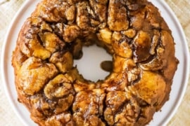 An untouched circular loaf of homemade monkey bread on a circular white plate next to a couple mugs of coffee.