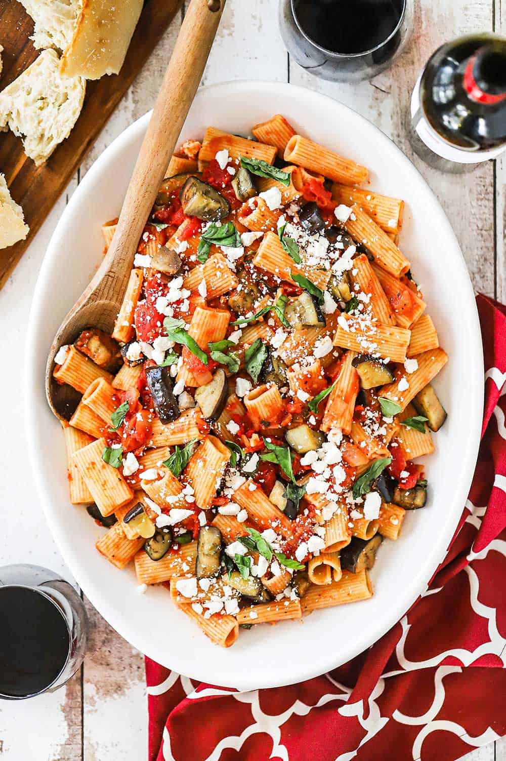 A large white oval serving platter filled with pasta alla Norma with a wooden spoon in the side all next to two glasses of red wine.