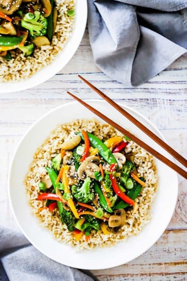 A white dinner plate filled with vegetable stir-fry over brown rice with two chop sticks sitting on the side.