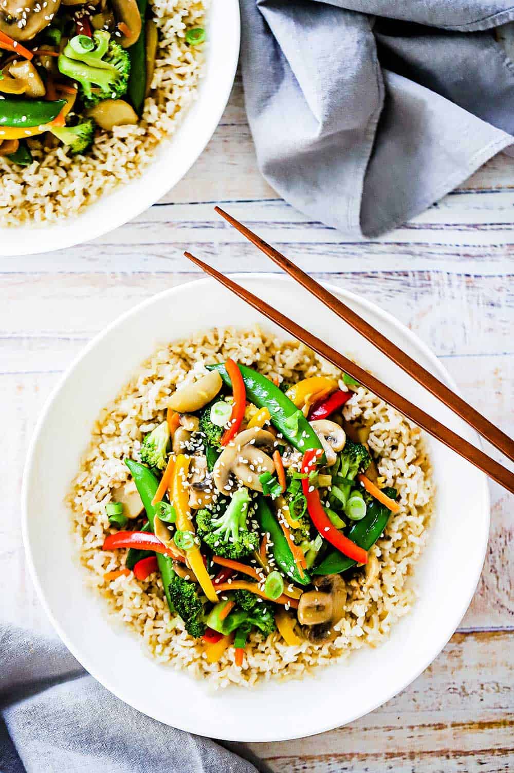 A white dinner plate filled with vegetable stir-fry over brown rice with two chop sticks sitting on the side. 