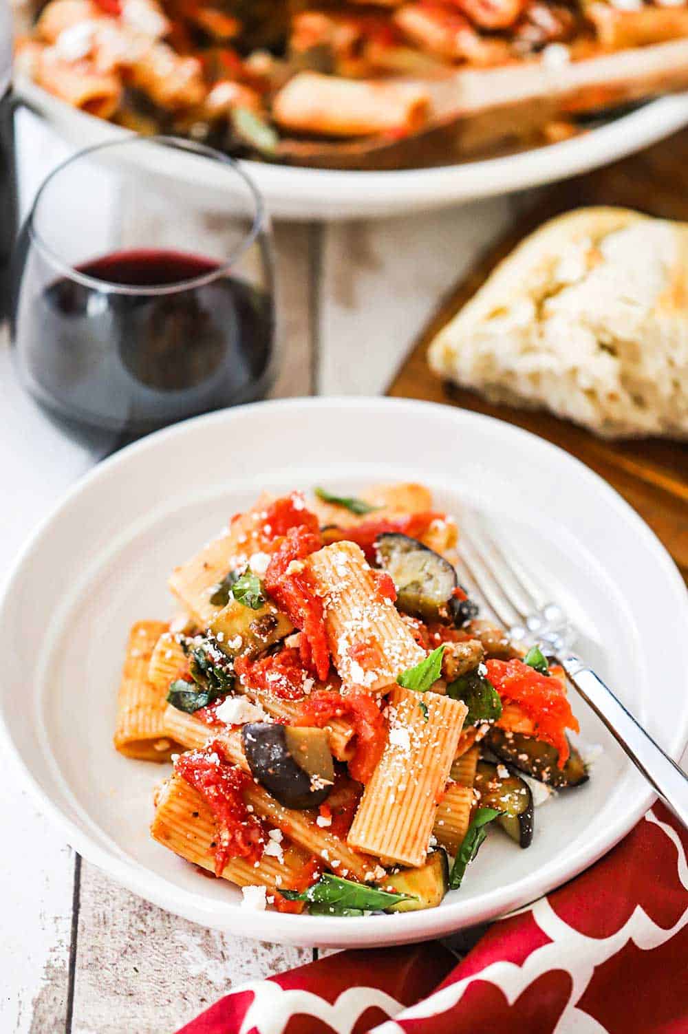 A circular white dinner pasta bowl filled with a serving of pasta alla Norma with a stemless glass of red wine nearby.