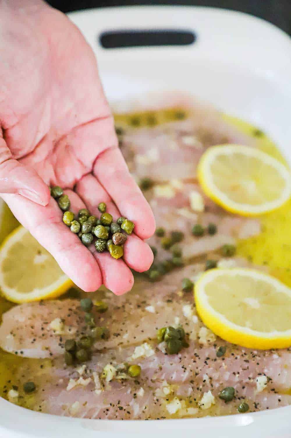 A hand dropping capers onto a filled with tilapia topped with lemon slices and minced garlic. 