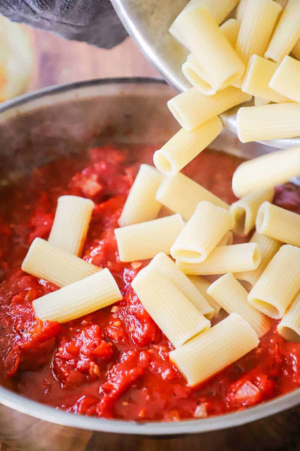 Cooked rigatoni pasta being transferred into a skillet filled with a simmering tomato sauce.