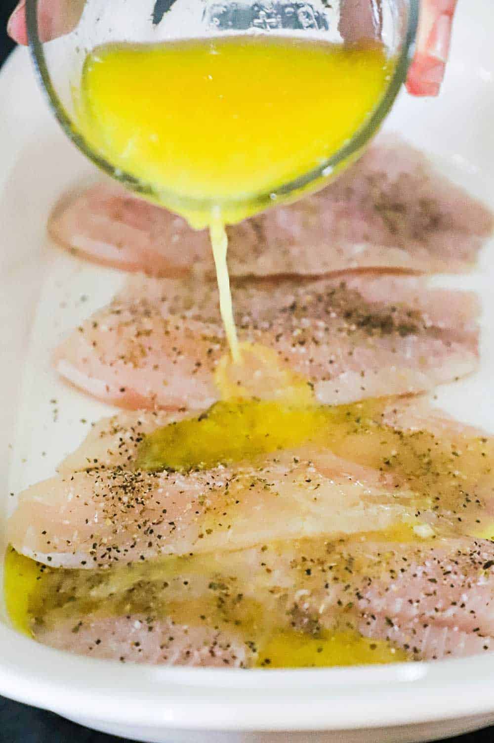 A person pouring a lemon butter sauce over tilapia fillets that have been seasoned with salt and pepper in a white baking dish. 