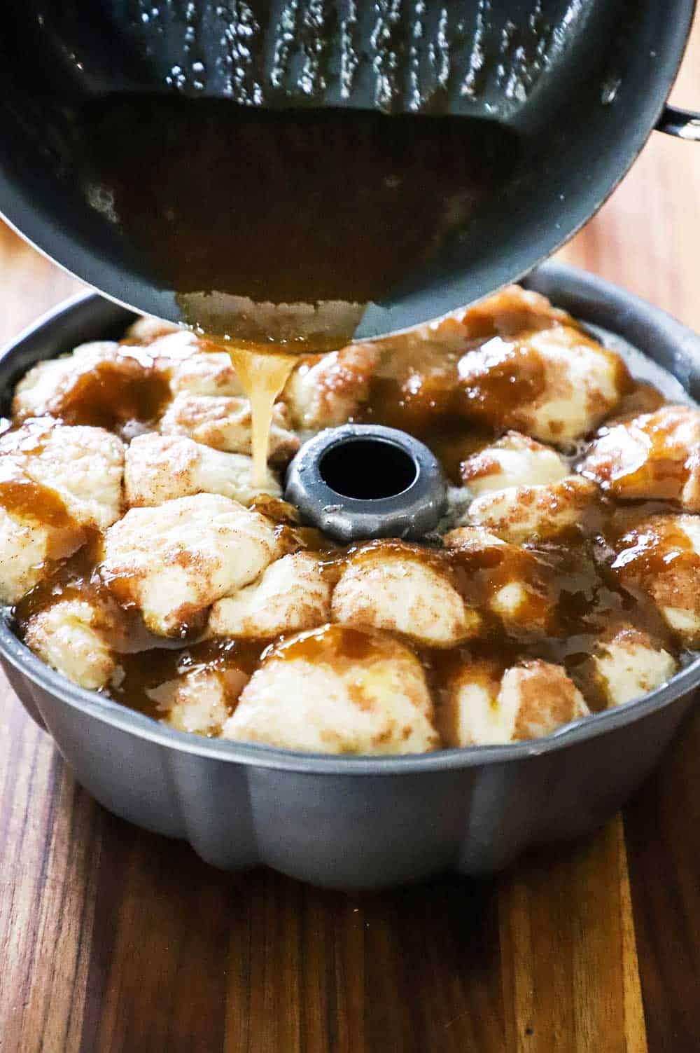 Caramel sauce being poured from a saucepan onto a bundt pan filled with risen balls of bread dough.