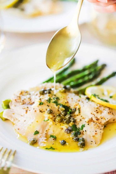 A gold spoon pouring a lemon butter sauce over a dish filled with baked tilapia.