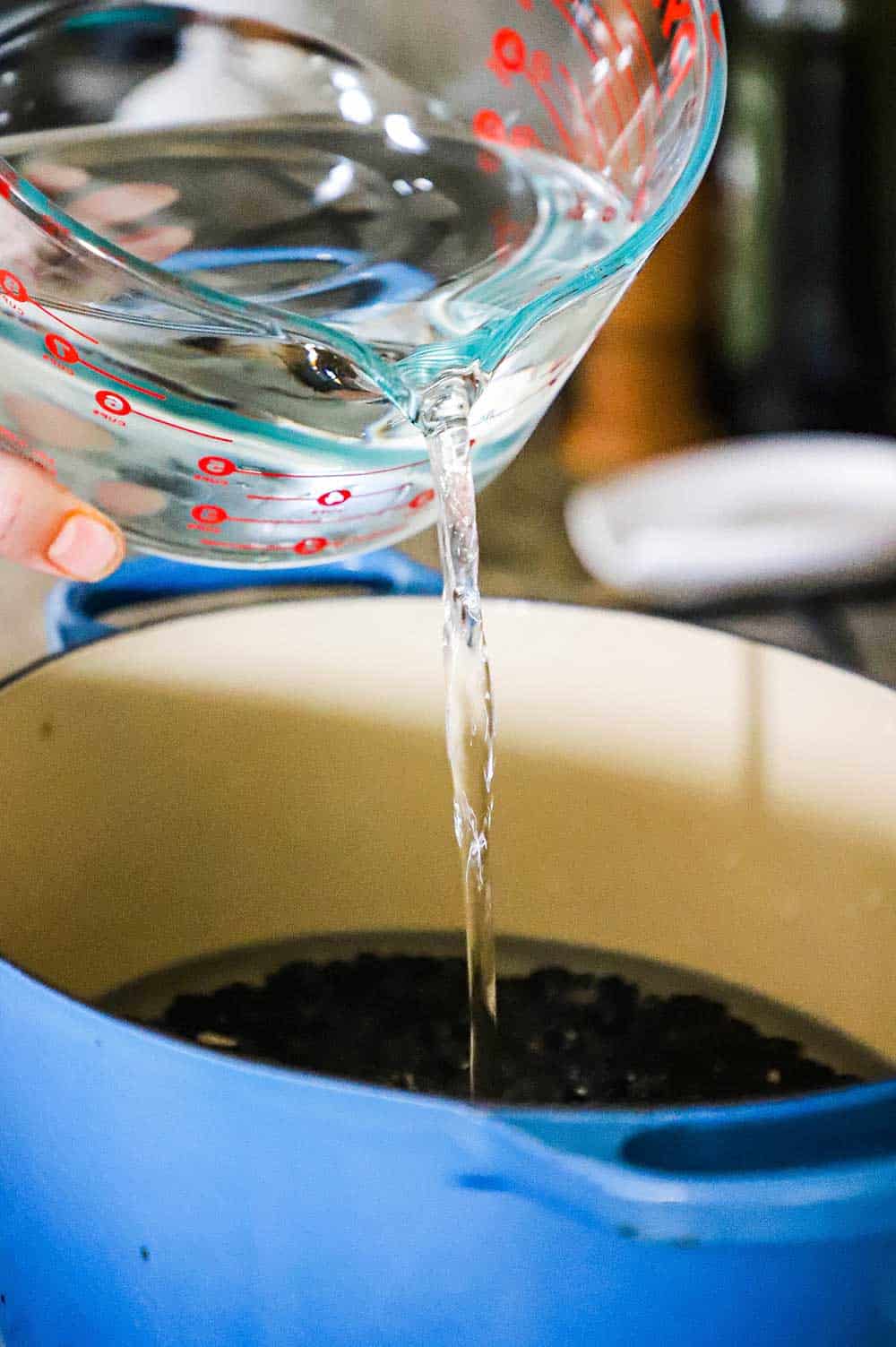 A person pouring water from an 8 cup measuring cup into a Dutch oven filled with dried black beans.