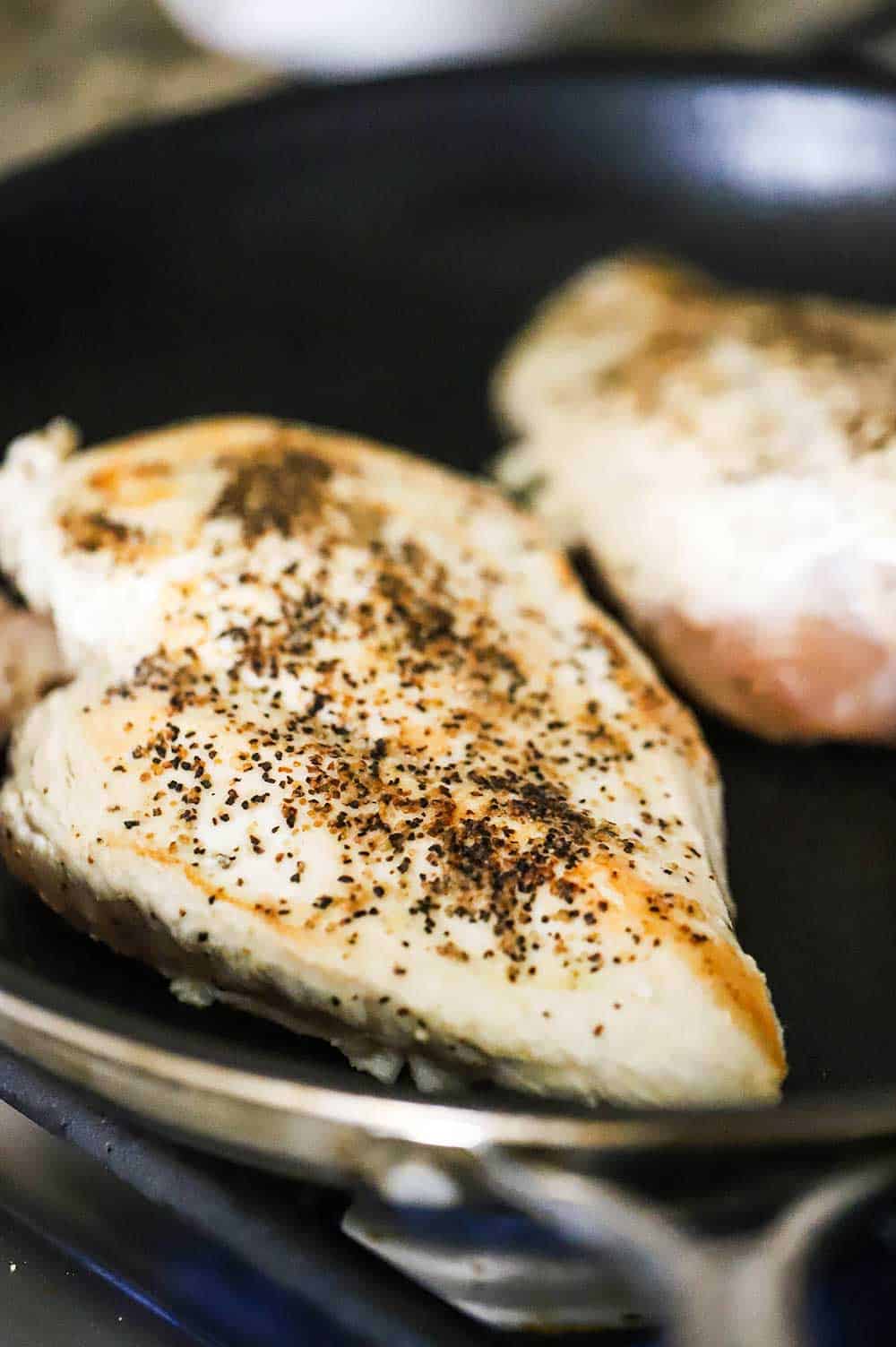 Two large chicken breasts that are being seared in a black non-stick skillet over a gas stove.
