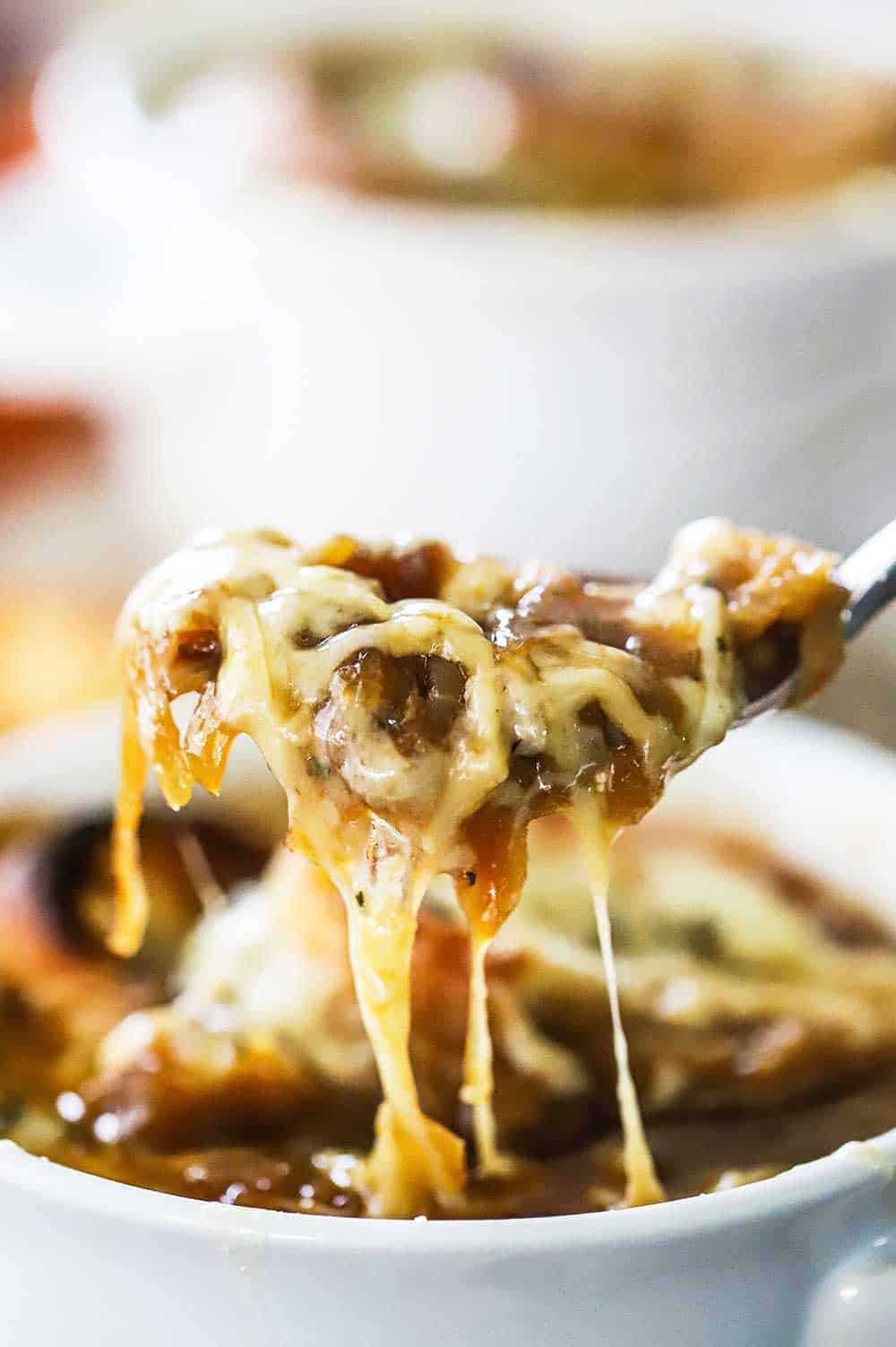 A spoon being raised out a bowl filled with French onion soup with cheese and onions dripping from the spoon.