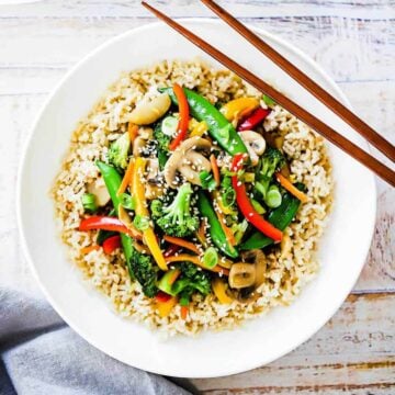 A white dinner plate filled with vegetable stir-fry over brown rice with two chop sticks sitting on the side.