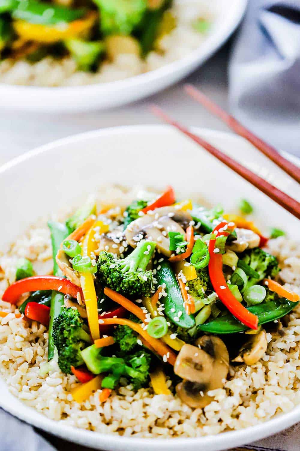 A white circular plate filled with vegetable stir-fry with brown chop sticks sitting on the side of the bowl. 