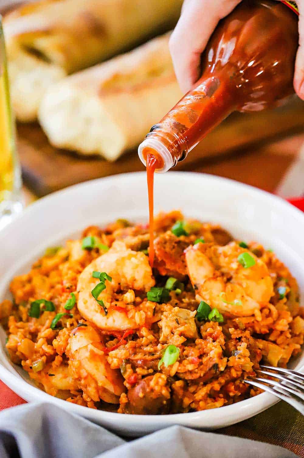 A person pouring hot sauce from a bottle onto a helping of jambalaya in a white bowl next to pieces of French bread.