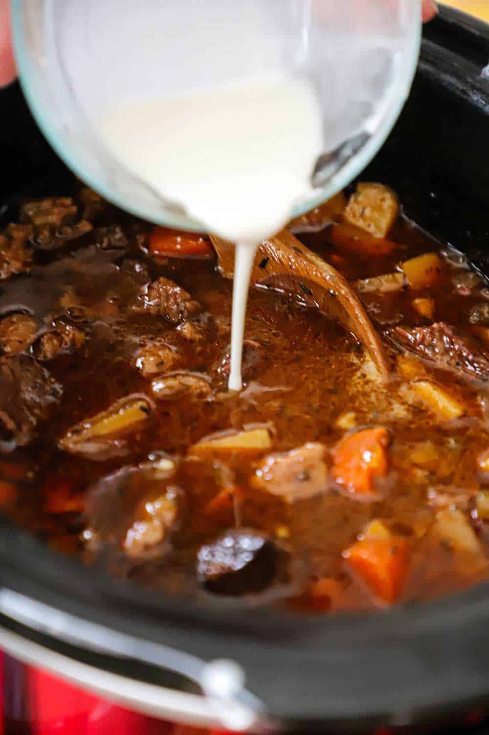 A cornstarch slurry being poured from a small glass bowl into a slow cooker filled with beef stew.