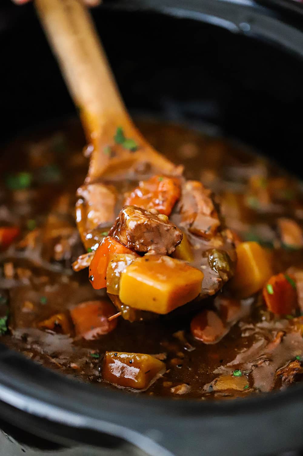 A wooden spoon being lifted out of a slow-cooker filled with beef stew.