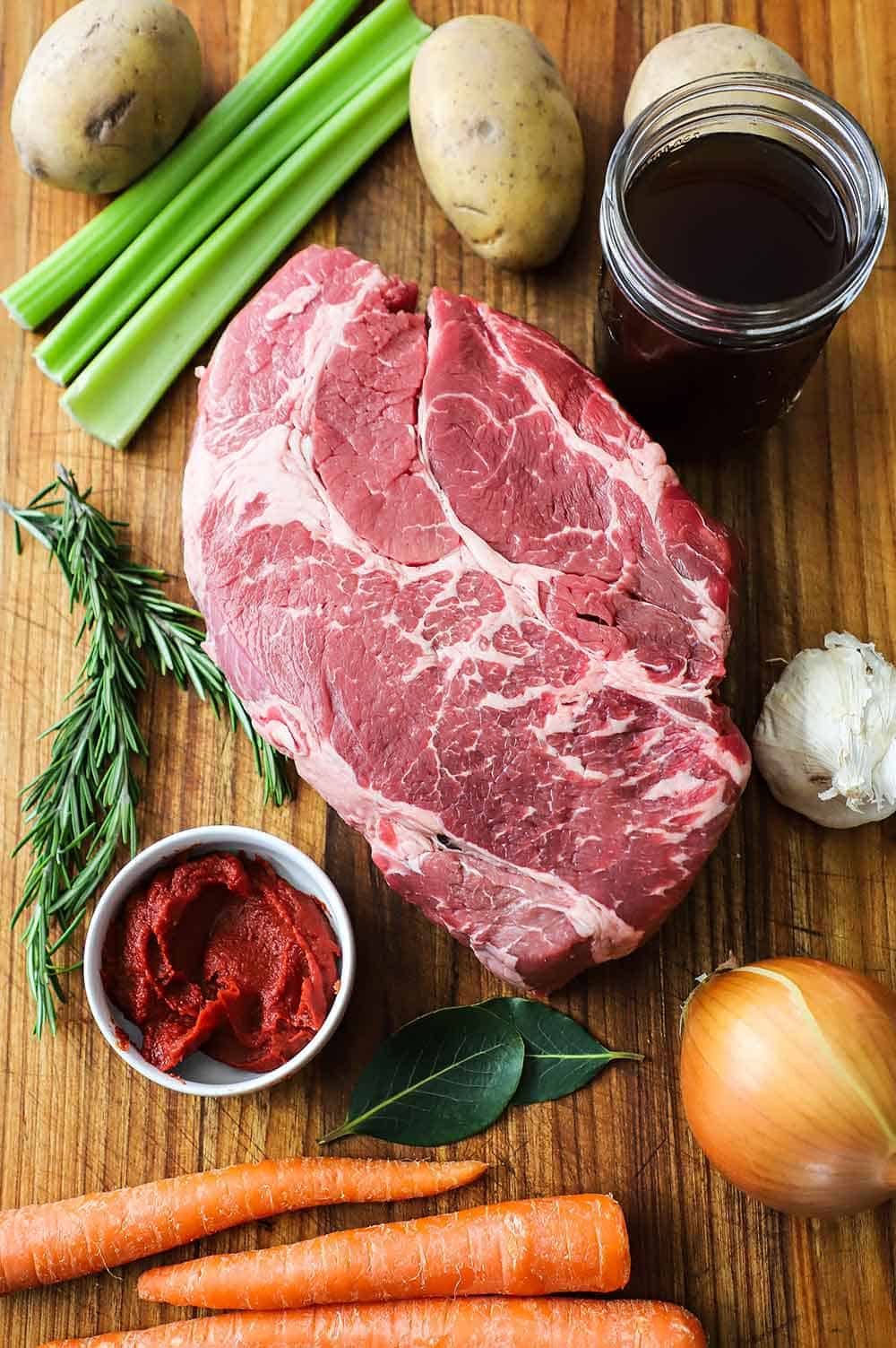 A wooden cutting board holding whole potatoes, celery stalks, beef broth in a jar, chuck roast, carrots, and on onion.