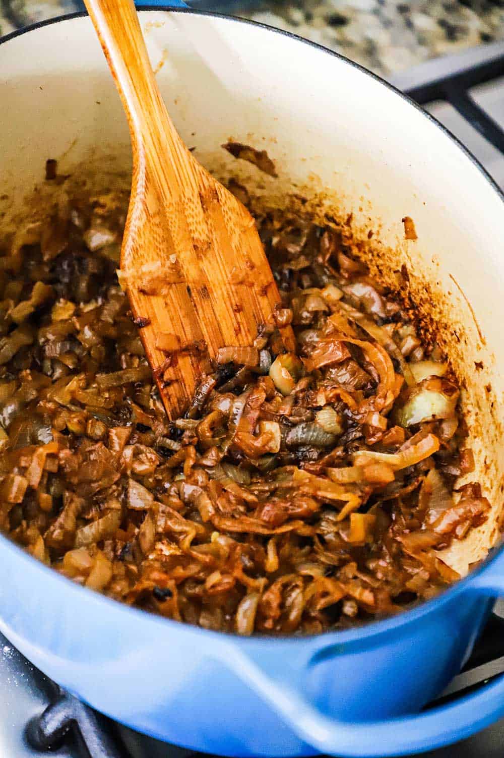 A large blue oval Dutch oven filled with caramelized onions with a wooden spatula inserted into the pot.