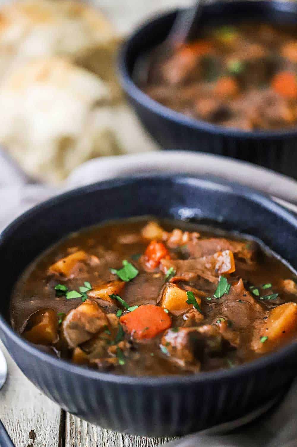 A blue soup bowl filled with slow-cooker beef stew with another bowl behind it sitting next to a loaf of bread.