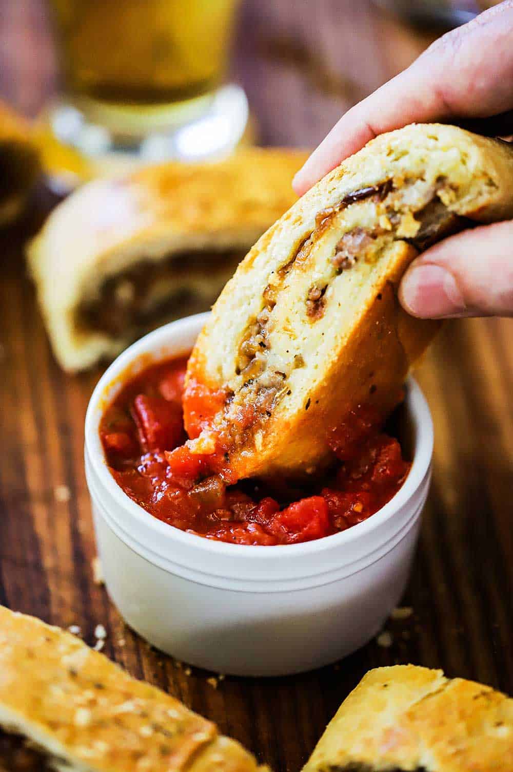 A person dipping a slice of sausage into a small white bowl of marinara sauce.
