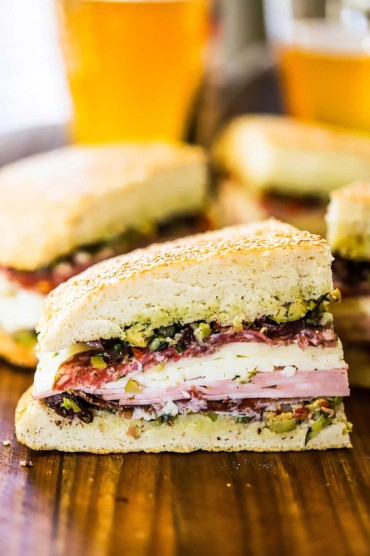 A quarter slice of a muffuletta sandwich on a cutting board surrounded by other slices and a mug of beer.