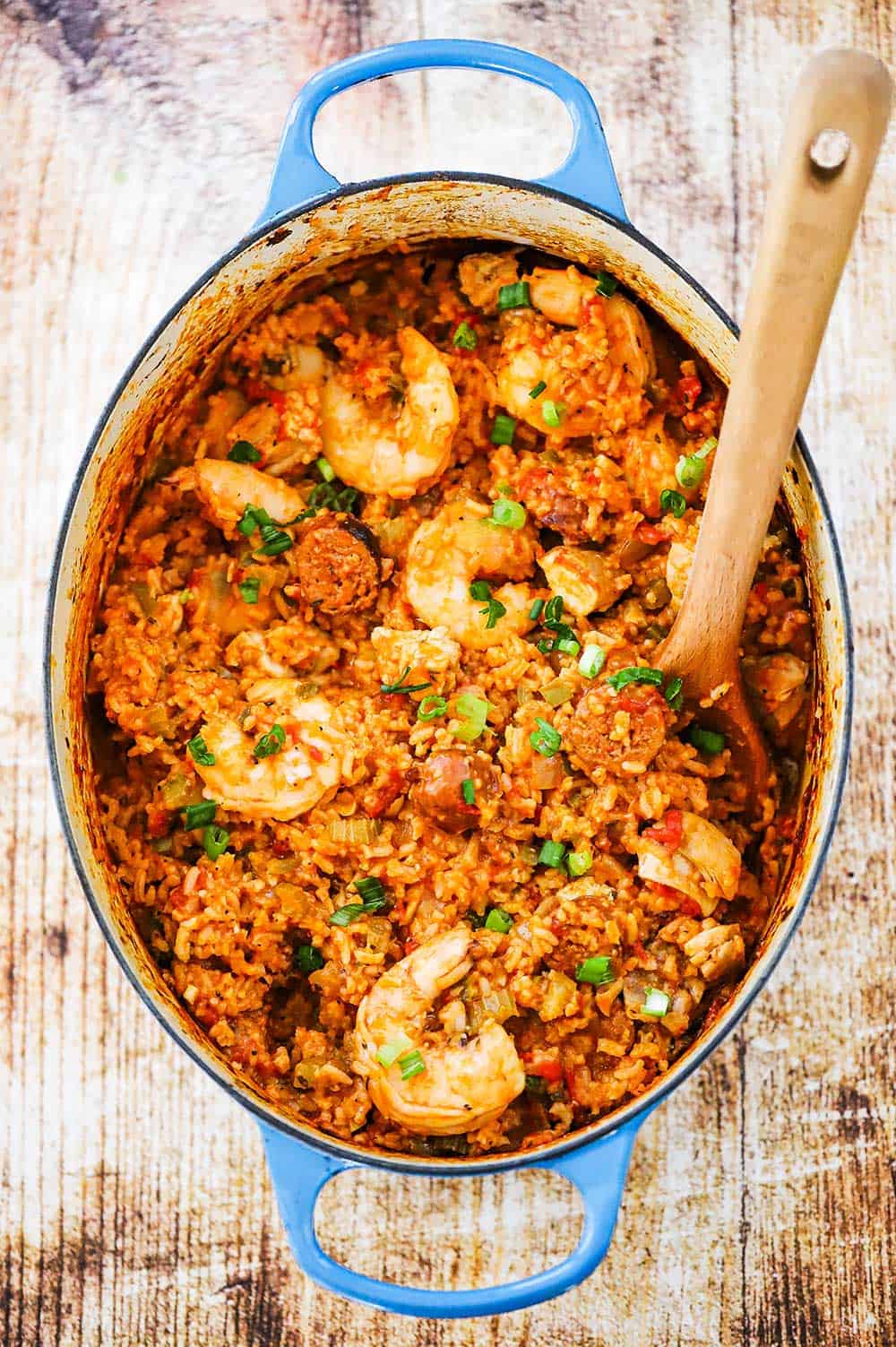 A large oval blue Dutch oven filled with jambalaya with a wooden spoon inserted into the mixture.