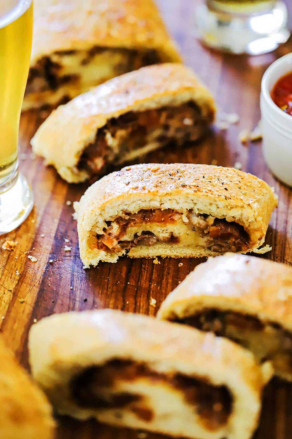 A line up of sliced sausage bread sitting on a wooden cutting board next to a glass of beer.