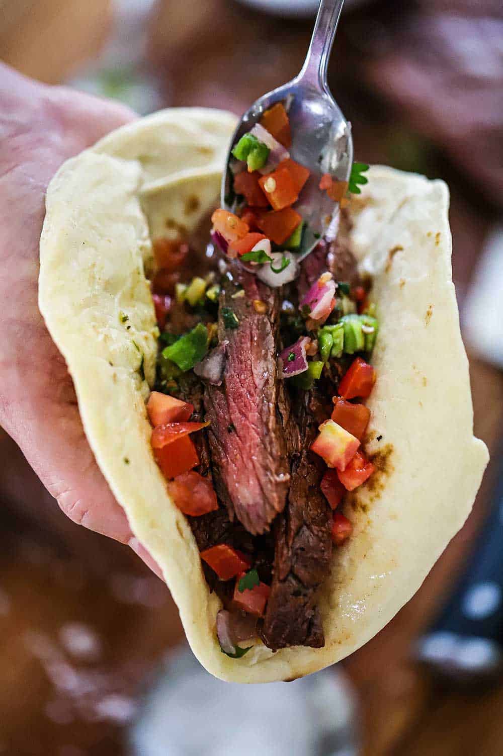 A person holding a flour tortilla that is filled with sliced carne asada and topped with pico de gallo.