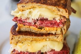 A grilled rueben sandwich that has been cut in half and then stacked on top of each other on a white plate with a pickle spear nearby.