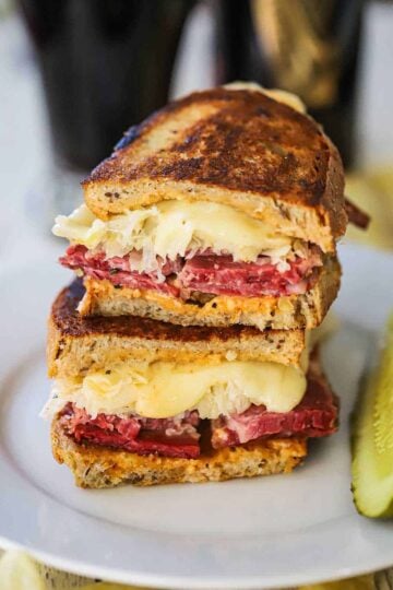 A grilled rueben sandwich that has been cut in half and then stacked on top of each other on a white plate with a pickle spear nearby.