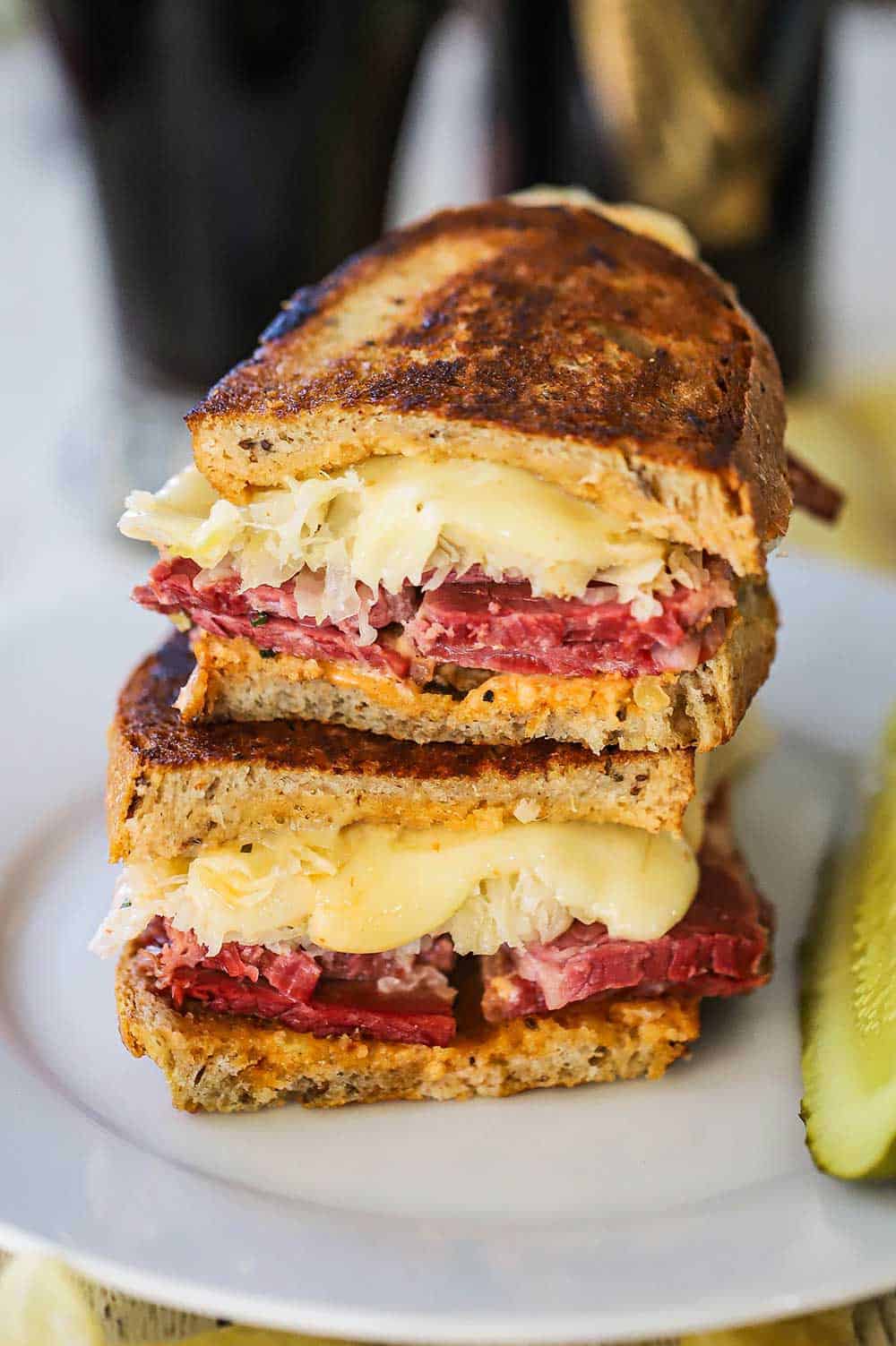 A grilled rueben sandwich that has been cut in half and then stacked on top of each other on a white plate with a pickle spear nearby.