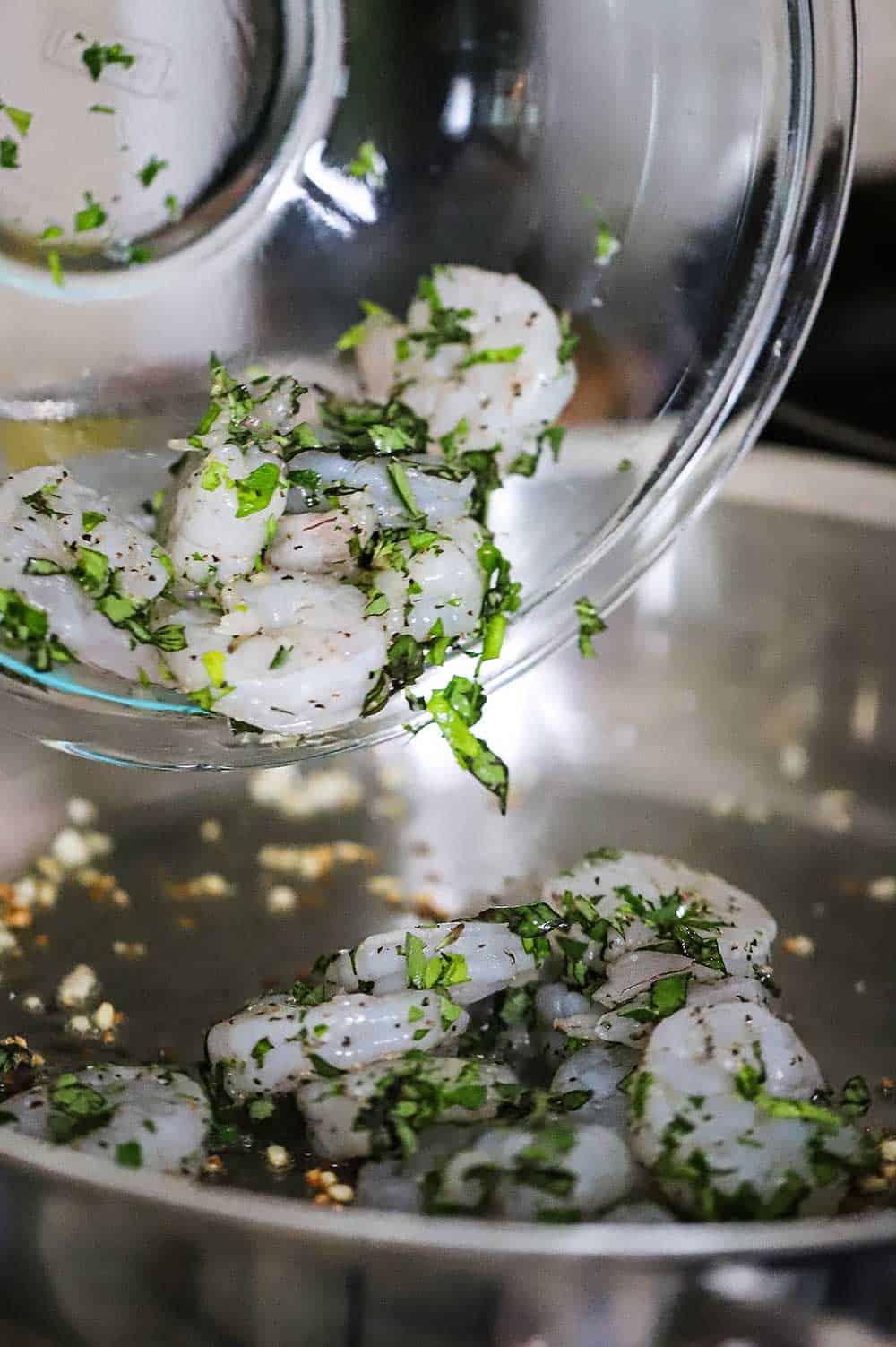 Uncooked shrimp that has been coated with fresh herbs being transferred into a skillet filled with sautéed garlic. 