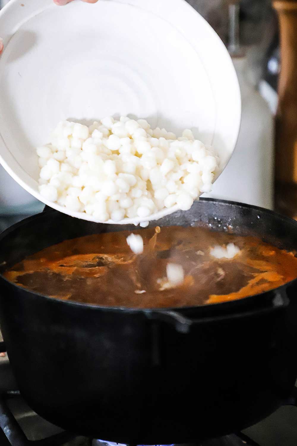 A person transferring white hominy from a white bowl into a black pot filled with pork broth.