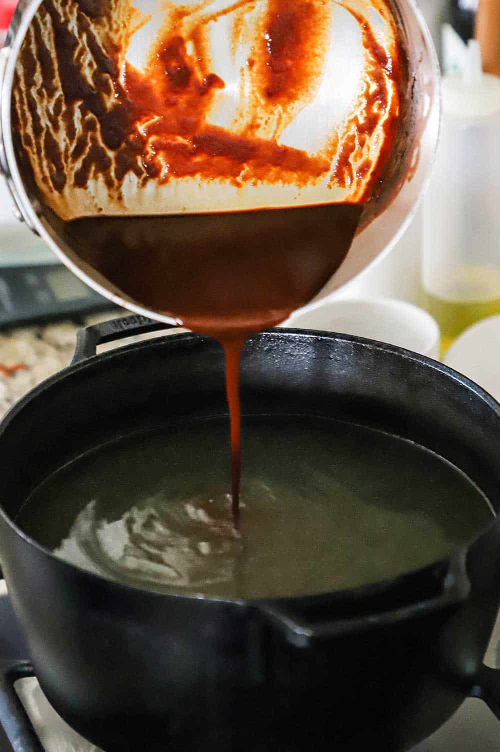 Ancho chili sauce being poured from a silver pot into a black pot filled with pork broth.