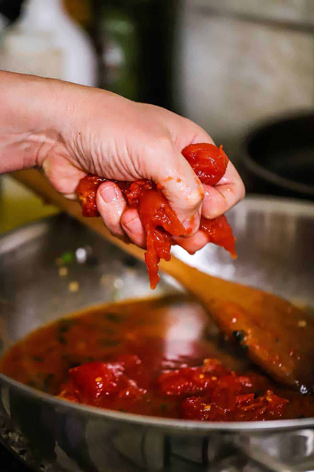 A person squeezing whole Italian tomatoes into a skillet filled with marinara sauce. 