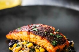 A slice of a blackened salmon fillet sitting on a corn and black bean salad on a black dinner plate.